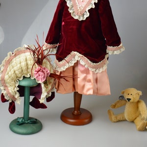 May include: A red velvet dress with a pink satin skirt and lace trim, a white and pink hat with a feather, and a brown teddy bear.