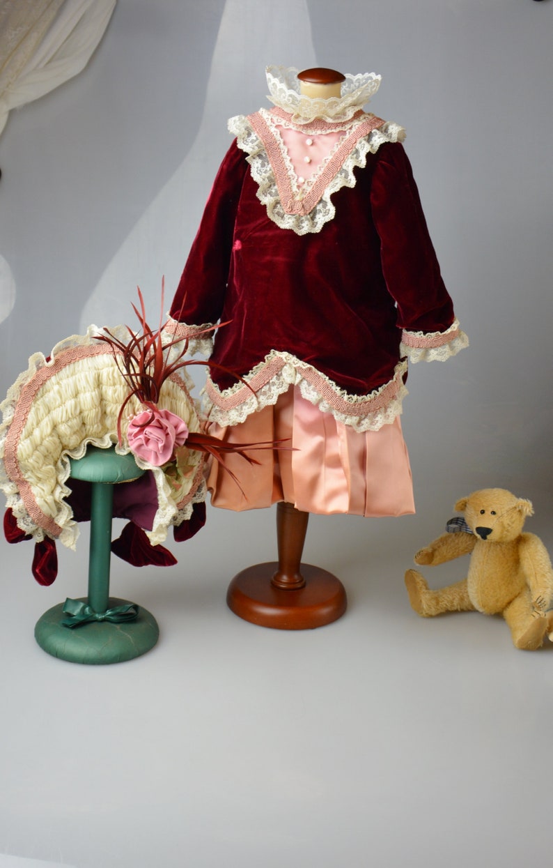 May include: A red velvet dress with a pink satin skirt and lace trim, a white and pink hat with a feather, and a brown teddy bear.