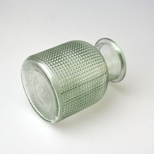 May include: A clear, light green glass vase with a cylindrical body featuring a textured, grid-like pattern across its exterior surface. The vase has a smooth, rounded neck and a wide, circular opening at the top, with a flat, circular base.