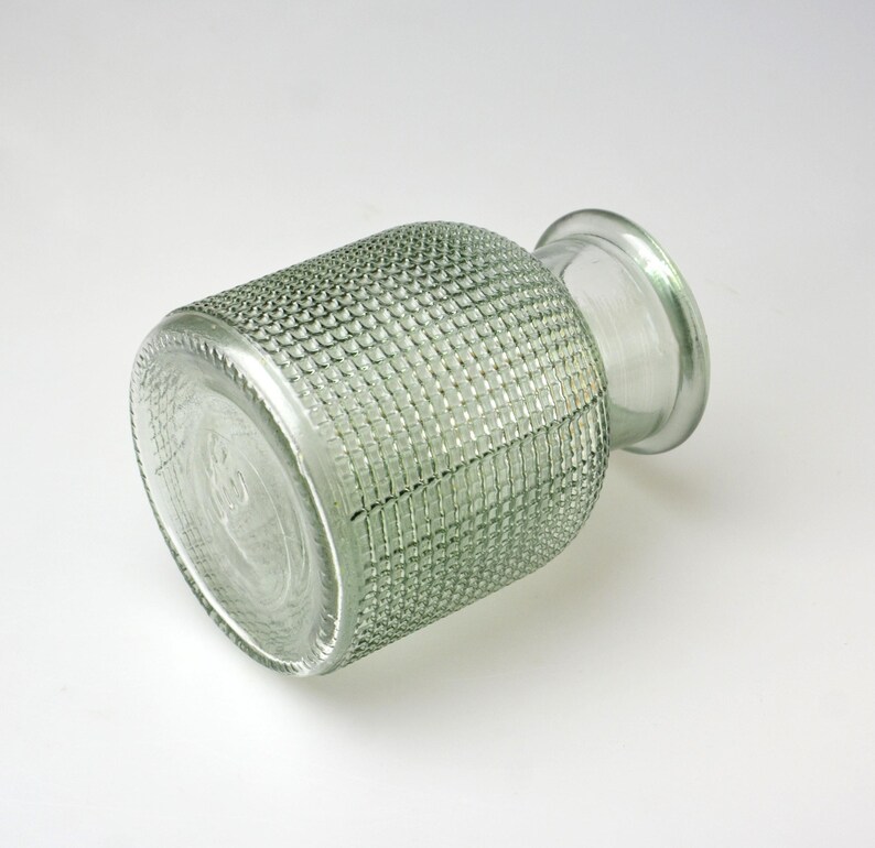 May include: A clear, light green glass vase with a cylindrical body featuring a textured, grid-like pattern across its exterior surface. The vase has a smooth, rounded neck and a wide, circular opening at the top, with a flat, circular base.