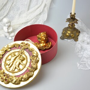 May include: A red round box with a gold lid. The lid has a raised design of two cherubs holding a ribbon. Inside the box is a gold cherub head and a pearl necklace.