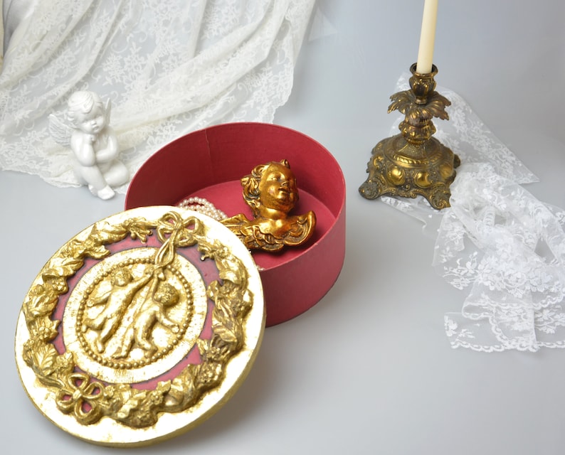 May include: A red round box with a gold lid. The lid has a raised design of two cherubs holding a ribbon. Inside the box is a gold cherub head and a pearl necklace.