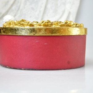 May include: A round, red, decorative box with a gold top. The top has a raised, ornate design.