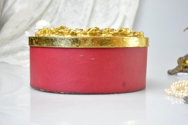 May include: A round, red, decorative box with a gold top. The top has a raised, ornate design.
