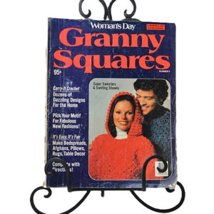 May include: A vintage Woman's Day Granny Squares magazine issue number 4, displayed on a black metal stand. The cover features bold red text on a dark blue background, showcasing a person in a red hooded crochet sweater next to another person.