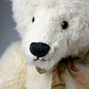May include: A white teddy bear with black eyes and a black nose. The bear is wearing a green and brown ribbon around its neck.