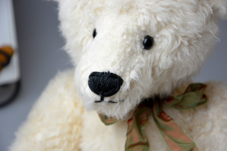 May include: A white teddy bear with black eyes and a black nose. The bear is wearing a green and brown ribbon around its neck.