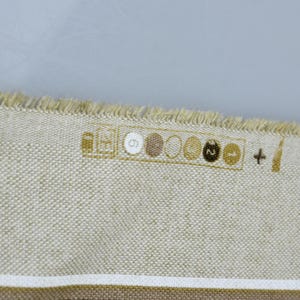 Puede incluir: Primer plano de una tela beige con textura tejida y borde deshilachado. La tela tiene una franja marr&oacute;n en la parte inferior y una serie de peque&ntilde;os dise&ntilde;os circulares en fila. Los dise&ntilde;os son en tonos marrones y beige.