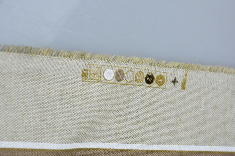 Puede incluir: Primer plano de una tela beige con textura tejida y borde deshilachado. La tela tiene una franja marr&oacute;n en la parte inferior y una serie de peque&ntilde;os dise&ntilde;os circulares en fila. Los dise&ntilde;os son en tonos marrones y beige.