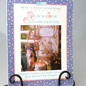 May include: A book titled "Quick-Sew Celebrations" on a black metal stand. The book cover has a blue floral pattern border and features a picture of a birthday scene with a cake and candles. The text reads "Decorative Creations for Festive Occasions".