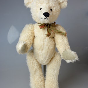 May include: A white teddy bear with black eyes and a black nose. The bear is wearing a green and brown ribbon around its neck. The bear is standing on a white stand.