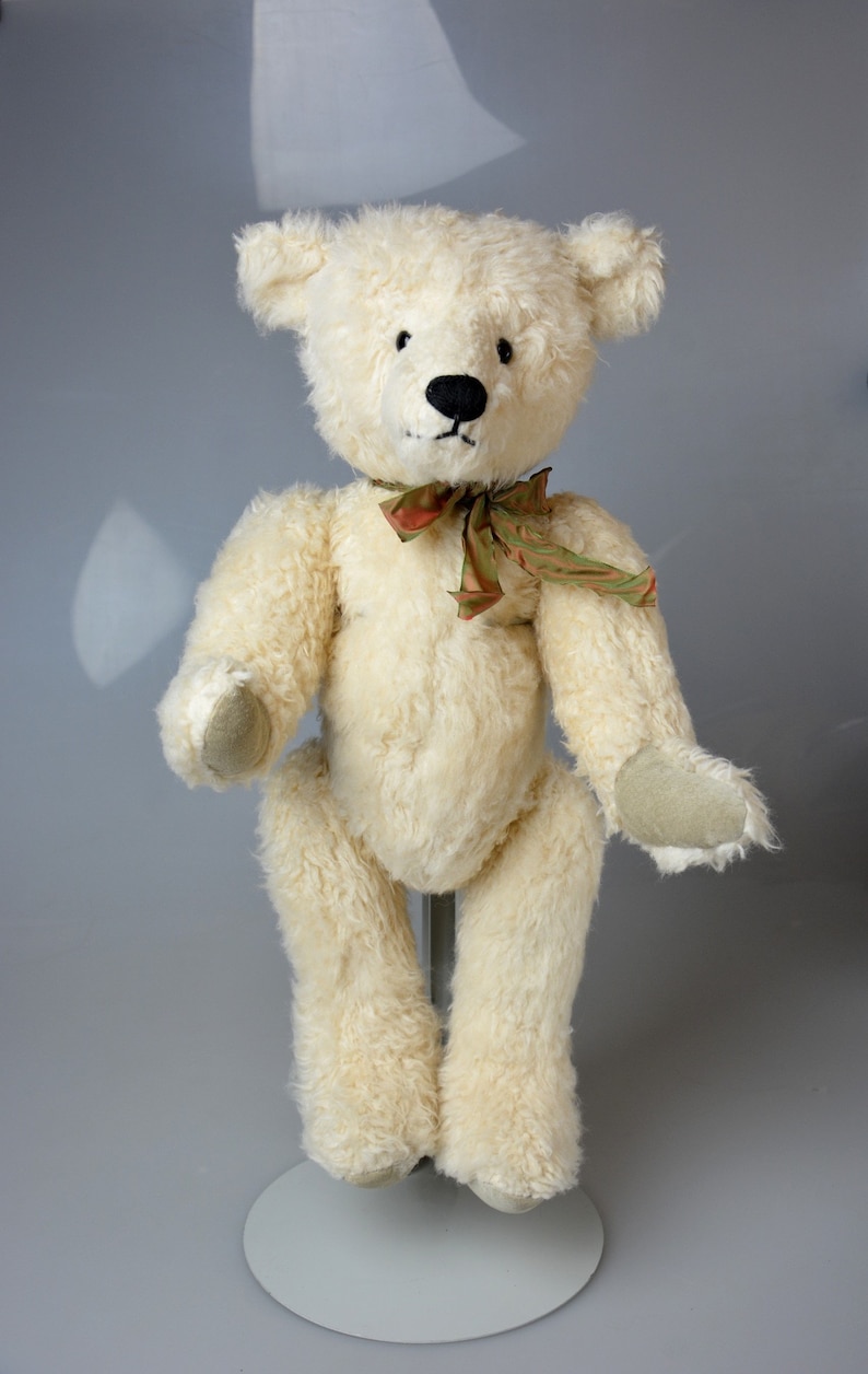 May include: A white teddy bear with black eyes and a black nose. The bear is wearing a green and brown ribbon around its neck. The bear is standing on a white stand.
