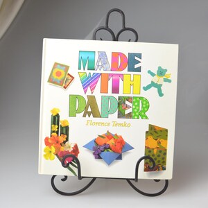 May include: A book titled "Made With Paper" by Florence Temko, featuring colorful paper crafts on the cover, displayed on a black metal book stand.