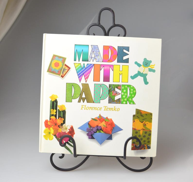 May include: A book titled "Made With Paper" by Florence Temko, featuring colorful paper crafts on the cover, displayed on a black metal book stand.