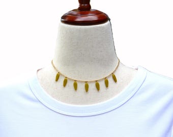 May include: A delicate gold-toned beaded choker necklace featuring seven evenly spaced, olive-green, teardrop-shaped charms hanging from the chain. The necklace is displayed on a neutral-colored mannequin wearing a plain white crew-neck t-shirt.