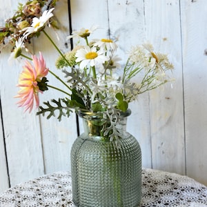 May include: A clear, sage green glass vase with a textured, grid-like pattern on the exterior. The vase contains a floral arrangement featuring white daisies with yellow centers, a pink and yellow dahlia, and silvery-green dusty miller foliage.