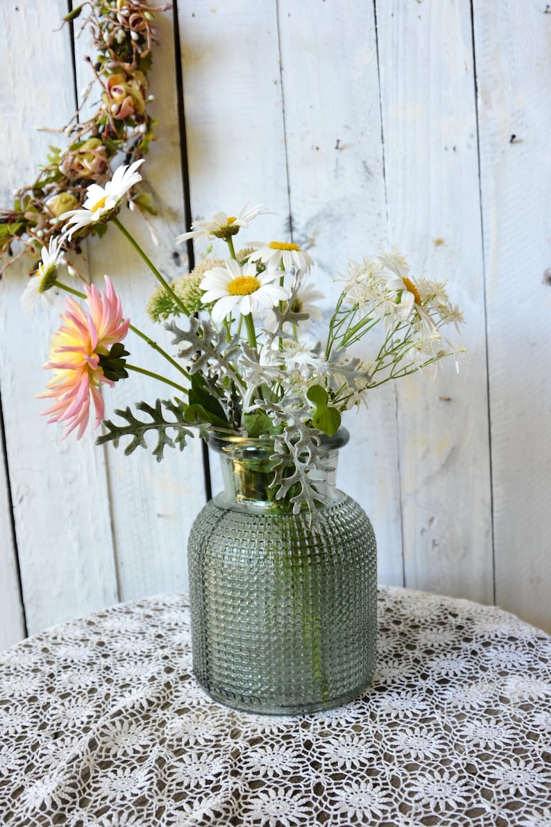 May include: A clear, sage green glass vase with a textured, grid-like pattern on the exterior. The vase contains a floral arrangement featuring white daisies with yellow centers, a pink and yellow dahlia, and silvery-green dusty miller foliage.