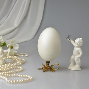 Puede incluir: Una figurita de cer&aacute;mica blanca de un querub&iacute;n sosteniendo un anillo, un huevo blanco sobre un soporte dorado con un anillo y una cadena de perlas blancas sobre una superficie blanca.