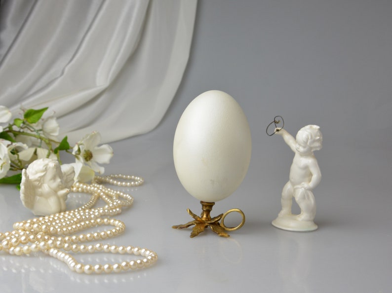 Puede incluir: Una figurita de cer&aacute;mica blanca de un querub&iacute;n sosteniendo un anillo, un huevo blanco sobre un soporte dorado con un anillo y una cadena de perlas blancas sobre una superficie blanca.