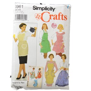 Könnte beinhalten: Eine Simplicity Crafts Schnittmusterhülle, Nummer 5961, mit Illustrationen verschiedener Schürzenmodelle. Die weiße Papierverpackung zeigt mehrere Schürzenstile in verschiedenen Farben und Mustern sowie den Schriftzug Simplicity Crafts.