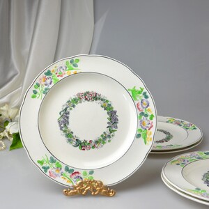 May include: A set of white ceramic plates with a floral wreath design in the center and around the scalloped edges. The plates have a black trim and are displayed on a gold-colored stand.