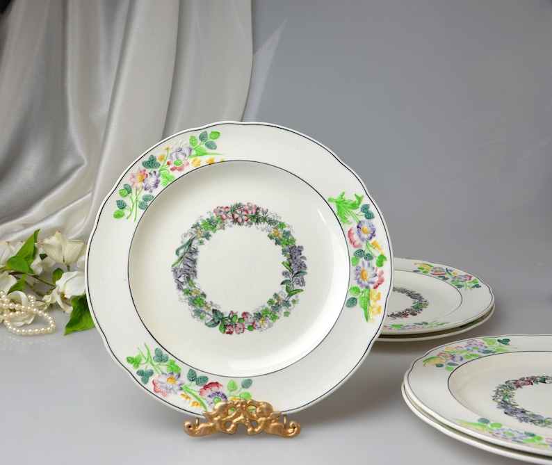 May include: A set of white ceramic plates with a floral wreath design in the center and around the scalloped edges. The plates have a black trim and are displayed on a gold-colored stand.