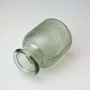 May include: A sage green glass vase with a textured hobnail pattern across its cylindrical body and a smooth, rounded rim. The vase is positioned at an angle, and a small cluster of white daisy-like flowers with yellow centers is visible.