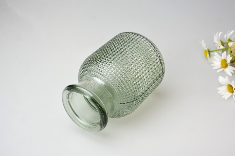 May include: A sage green glass vase with a textured hobnail pattern across its cylindrical body and a smooth, rounded rim. The vase is positioned at an angle, and a small cluster of white daisy-like flowers with yellow centers is visible.