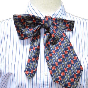 May include: A white button-down shirt with thin blue and red vertical stripes, featuring a large, tied neck scarf. The scarf is navy blue with a repeating geometric pattern of red rectangles and gold diamond shapes containing blue floral motifs.
