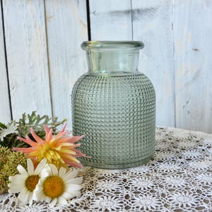 May include: A sage green glass vase with a wide, rounded body and a narrow neck. The exterior surface features a textured, grid-like pattern of small raised squares. The vase has a thick, smooth rim at the top and a clear, translucent appearance.