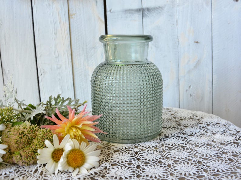 May include: A sage green glass vase with a wide, rounded body and a narrow neck. The exterior surface features a textured, grid-like pattern of small raised squares. The vase has a thick, smooth rim at the top and a clear, translucent appearance.