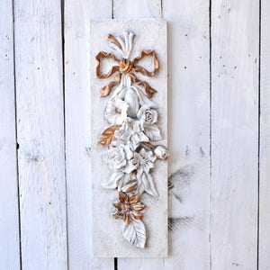 May include: A white and gold floral wall decor with a bow at the top. The flowers are sculpted and have a 3D effect. The piece is mounted on a white background.