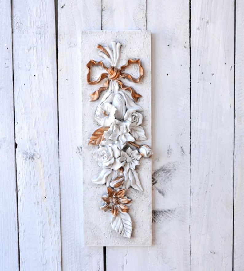 May include: A white and gold floral wall decor with a bow at the top. The flowers are sculpted and have a 3D effect. The piece is mounted on a white background.