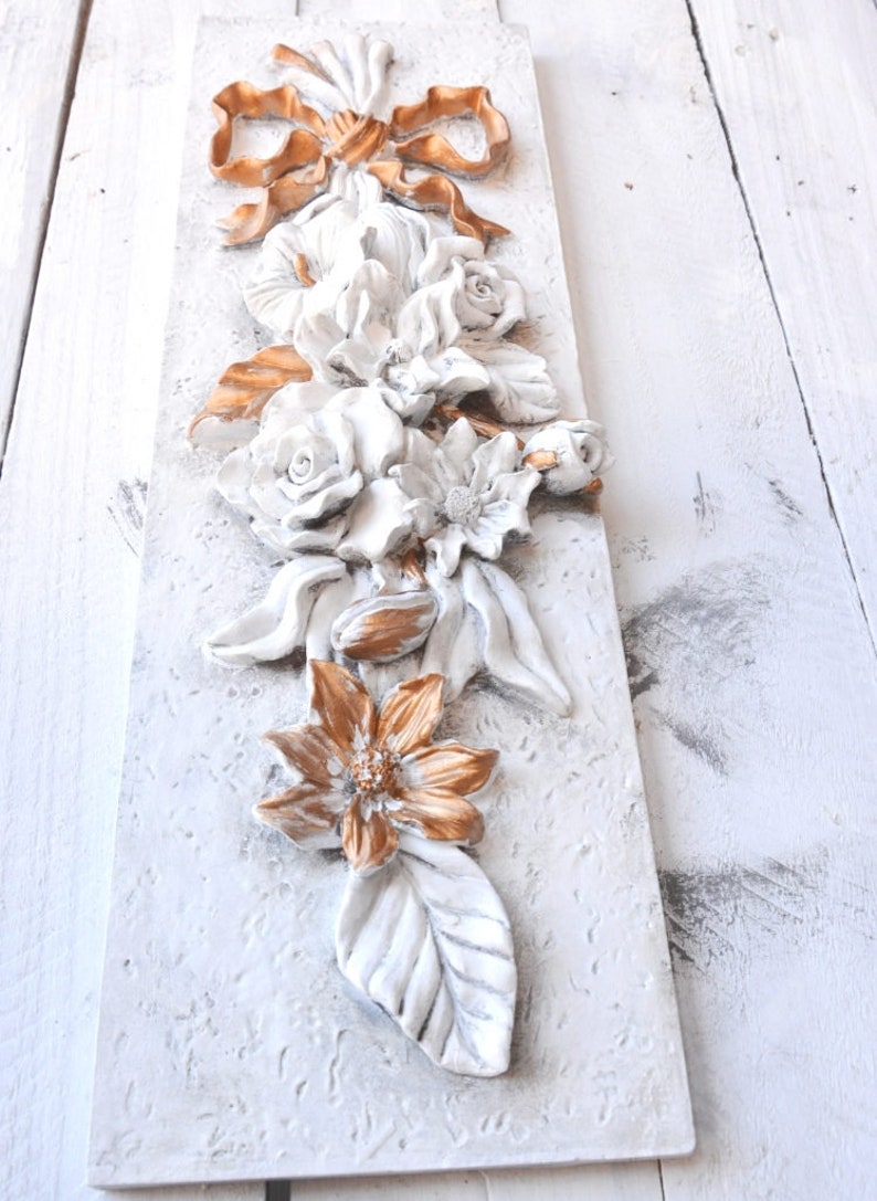 May include: A white and gold floral wall decor with a 3D design. The flowers are made of plaster and painted in a white and gold color scheme. The flowers are arranged in a cascading design, with a bow at the top.