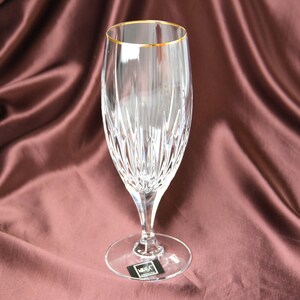 Pu&ograve; includere: Un calice da champagne trasparente con bordo dorato, con un design in cristallo tagliato. Lo stelo e la base sono anch'essi trasparenti, su uno sfondo di tessuto drappeggiato marrone rossiccio. Il bicchiere &egrave; alto circa 23 cm.