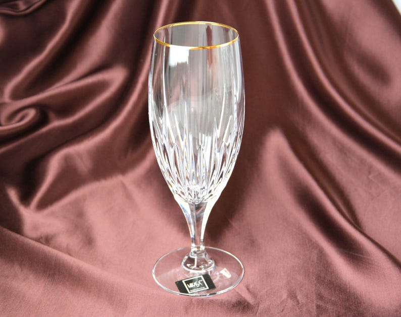 Pu&ograve; includere: Un calice da champagne trasparente con bordo dorato, con un design in cristallo tagliato. Lo stelo e la base sono anch'essi trasparenti, su uno sfondo di tessuto drappeggiato marrone rossiccio. Il bicchiere &egrave; alto circa 23 cm.