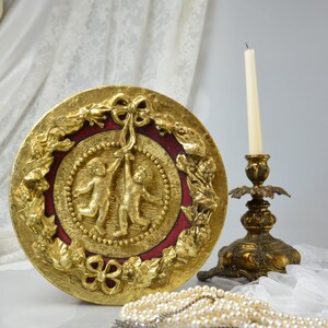 May include: A round, gold-leafed decorative wall plaque with a red center and two cherubs holding a ribbon. The plaque is surrounded by a wreath of leaves and flowers.