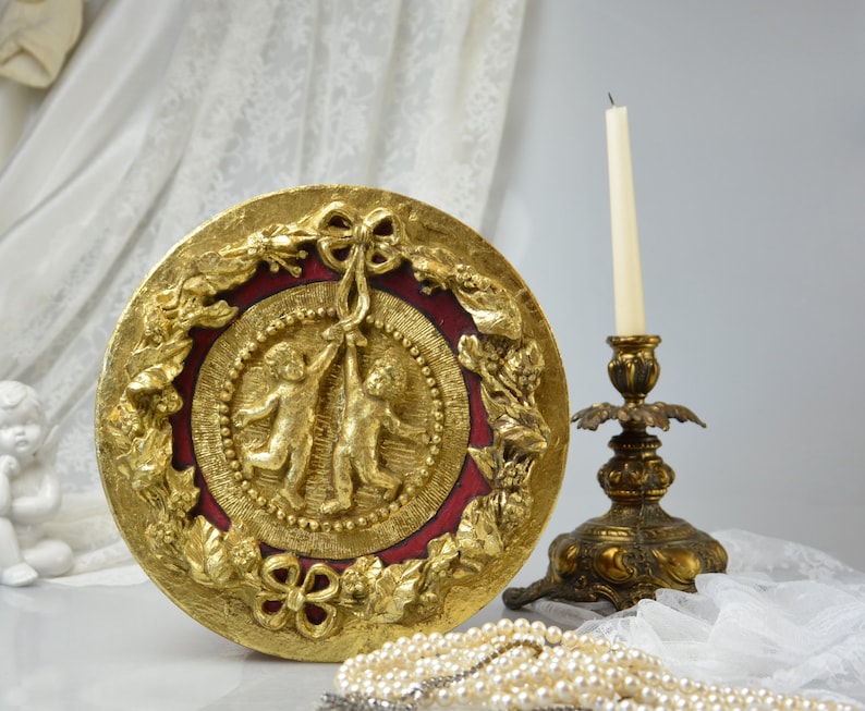 May include: A round, gold-leafed decorative wall plaque with a red center and two cherubs holding a ribbon. The plaque is surrounded by a wreath of leaves and flowers.