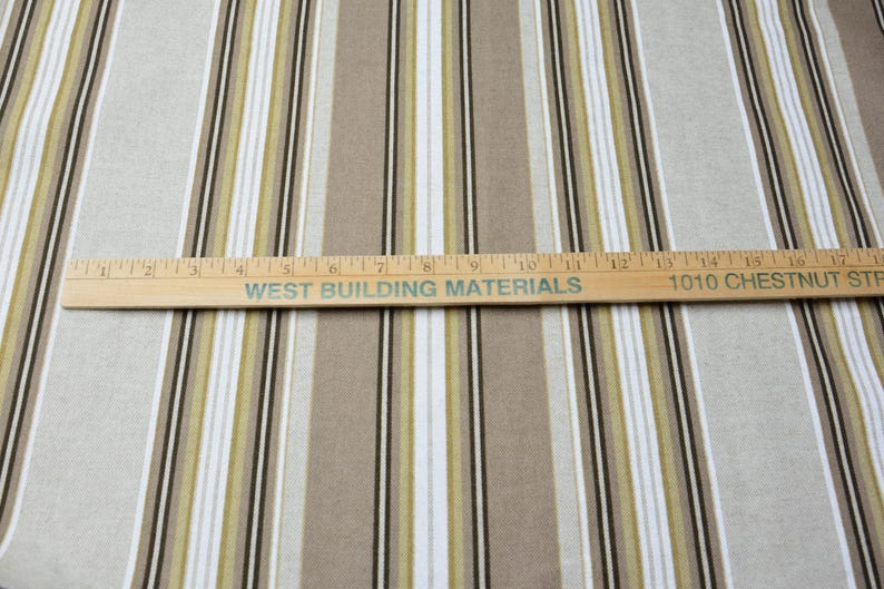 Puede incluir: Tela con rayas verticales en beige, blanco, verde oliva y negro. Una regla de madera con el texto "WEST BUILDING MATERIALS" y "1010 CHESTNUT ST" cruza la tela. La tela es adecuada para decoraci&oacute;n del hogar o ropa.