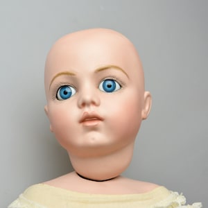 May include: A porcelain doll head with blue eyes and painted eyebrows. The doll has a light pink complexion and is wearing a white dress.