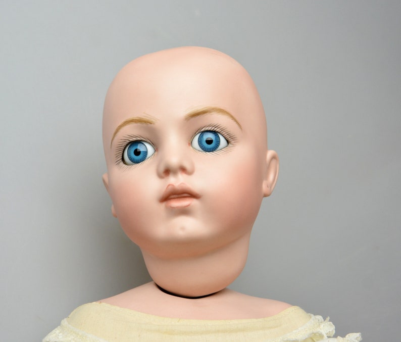 May include: A porcelain doll head with blue eyes and painted eyebrows. The doll has a light pink complexion and is wearing a white dress.