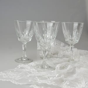 May include: Four clear crystal wine glasses with etched leaf designs. The glasses are displayed on a white lace tablecloth, creating an elegant and classic aesthetic. The stemware is ideal for serving wine.
