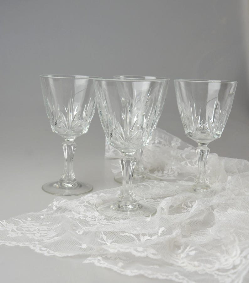 May include: Four clear crystal wine glasses with etched leaf designs. The glasses are displayed on a white lace tablecloth, creating an elegant and classic aesthetic. The stemware is ideal for serving wine.