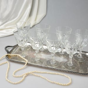 May include: A collection of eight clear crystal wine glasses with intricate starburst patterns, arranged on a silver serving tray. A strand of pearls is draped across the tray. The glasses are stemware.