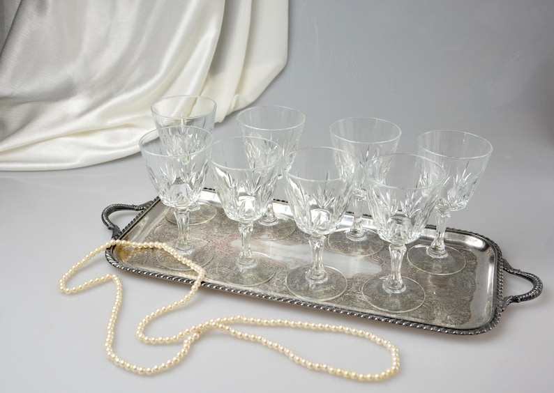 May include: A collection of eight clear crystal wine glasses with intricate starburst patterns, arranged on a silver serving tray. A strand of pearls is draped across the tray. The glasses are stemware.