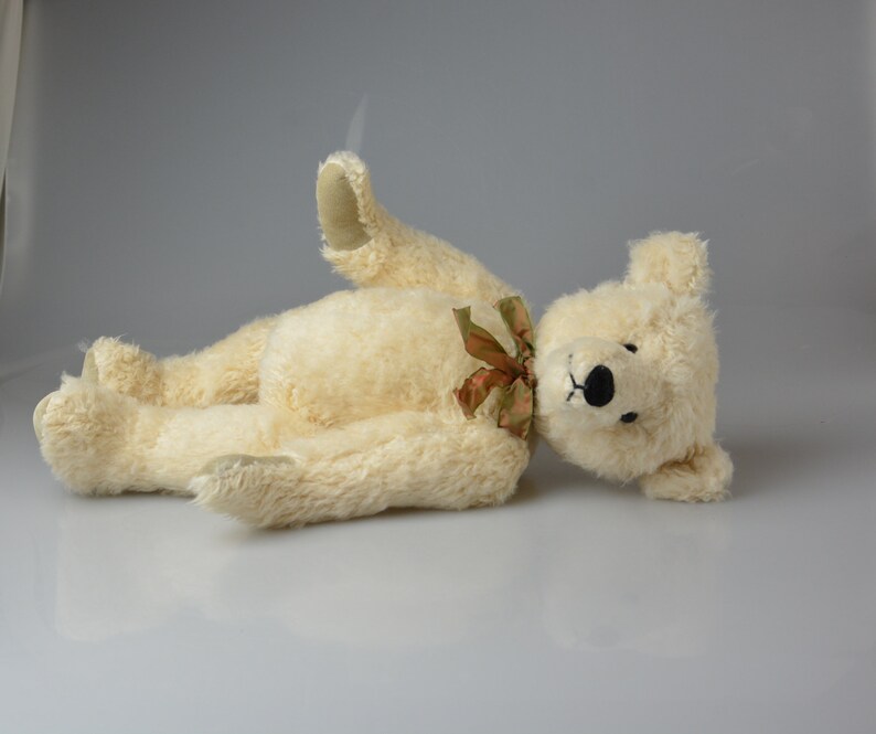 May include: A cream-colored teddy bear with a brown ribbon around its neck is lying on its back with its head turned to the side. The bear has black eyes and a black nose.