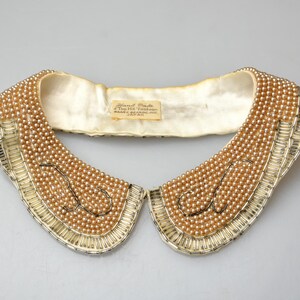 May include: A white satin collar with a beaded design. The beads are a light brown color and are arranged in a pattern of overlapping circles. The collar is trimmed with a gold-colored metal trim.