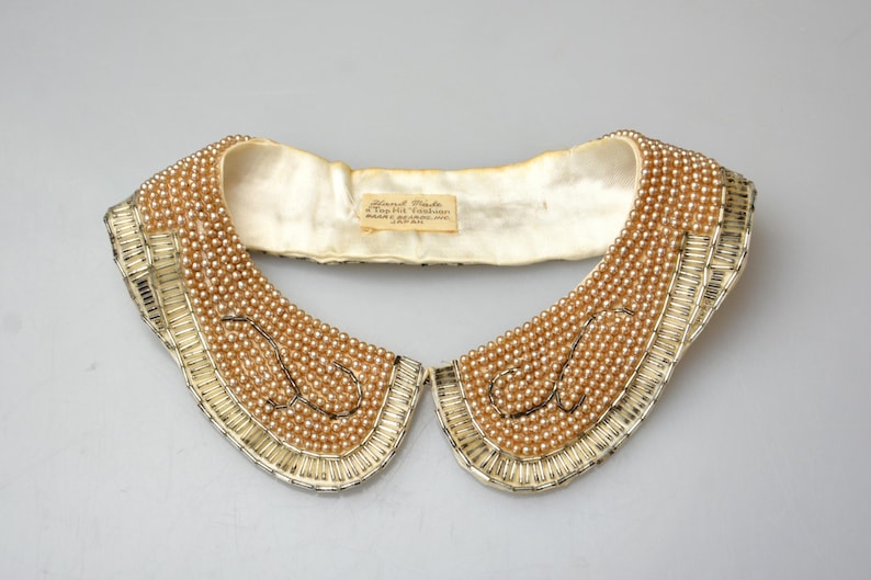 May include: A white satin collar with a beaded design. The beads are a light brown color and are arranged in a pattern of overlapping circles. The collar is trimmed with a gold-colored metal trim.