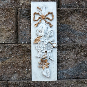 May include: White rectangular wall plaque with a raised floral design. The flowers are white with gold accents and a gold bow at the top.