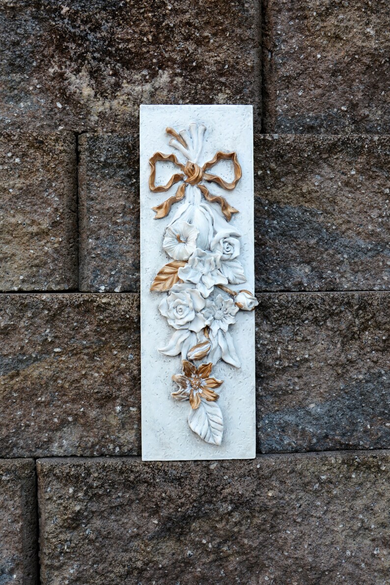 May include: White rectangular wall plaque with a raised floral design. The flowers are white with gold accents and a gold bow at the top.